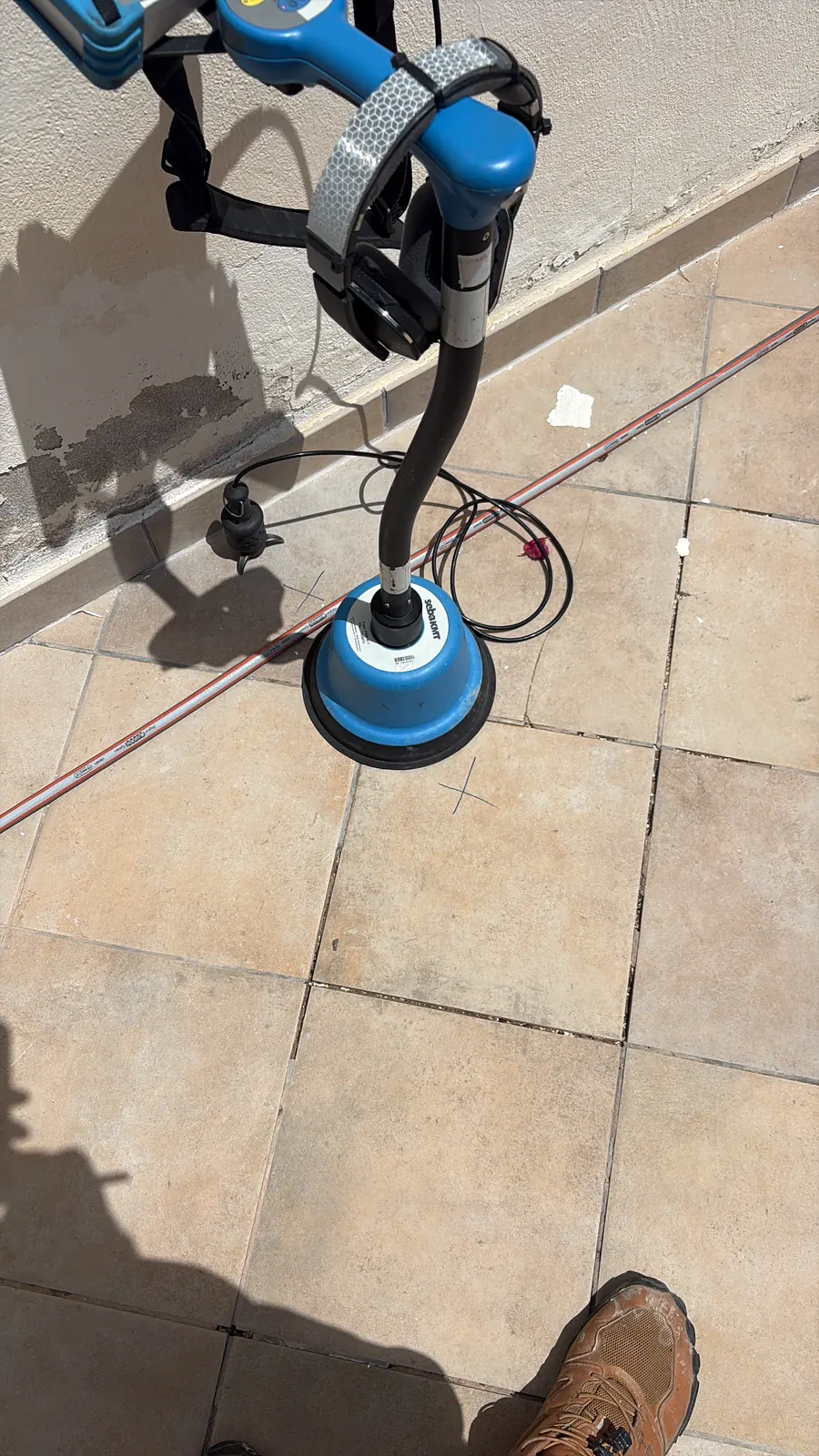 Sensor de suelo sebaKMT sobre una baldosa exterior, con cruces marcadas a lápiz señalando los puntos de escucha
